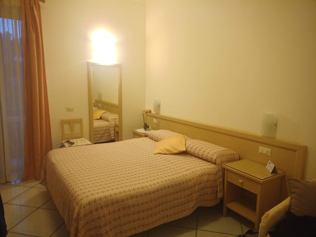 A hotel bedroom with a double bed, wooden furniture, and warm beige tones, featuring a mirror and bedside table.