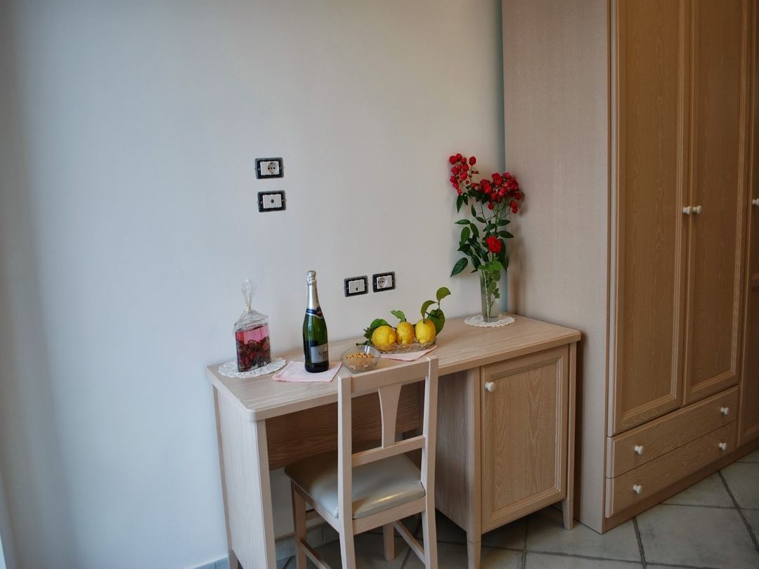 A wooden desk with a decorative arrangement featuring red flowers, lemons, champagne, and a beverage dispenser in a bright room.