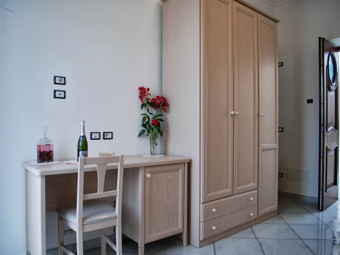 A bright bedroom corner with light wood furniture, including a desk with chair, wardrobe, fresh red flowers in a vase, and champagne bottle.